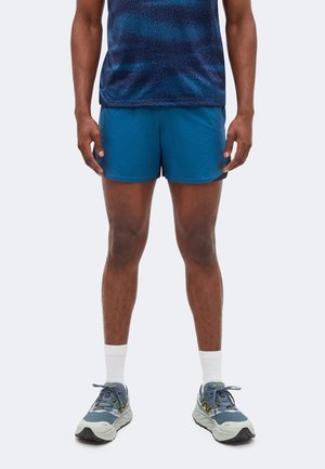 Mann mit blauen Sportshorts, blau gemustertem Hemd, weißen Crew-Socken und blauen Laufschuhen, stehend vor weißem Hintergrund.