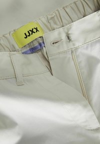 Bež hlače imaju gumirani pojas, patentni zatvarač, metalnu kopču i svijetlu žutu oznaku sa natpisom "JJXX" ispisanim u crnoj boji.