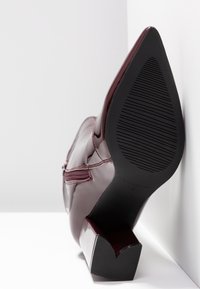 Botte à talon haut en cuir verni bordeaux à bout pointu, avec fermeture éclair latérale, semelle inclinée montrant un dessous en caoutchouc noir texturé.