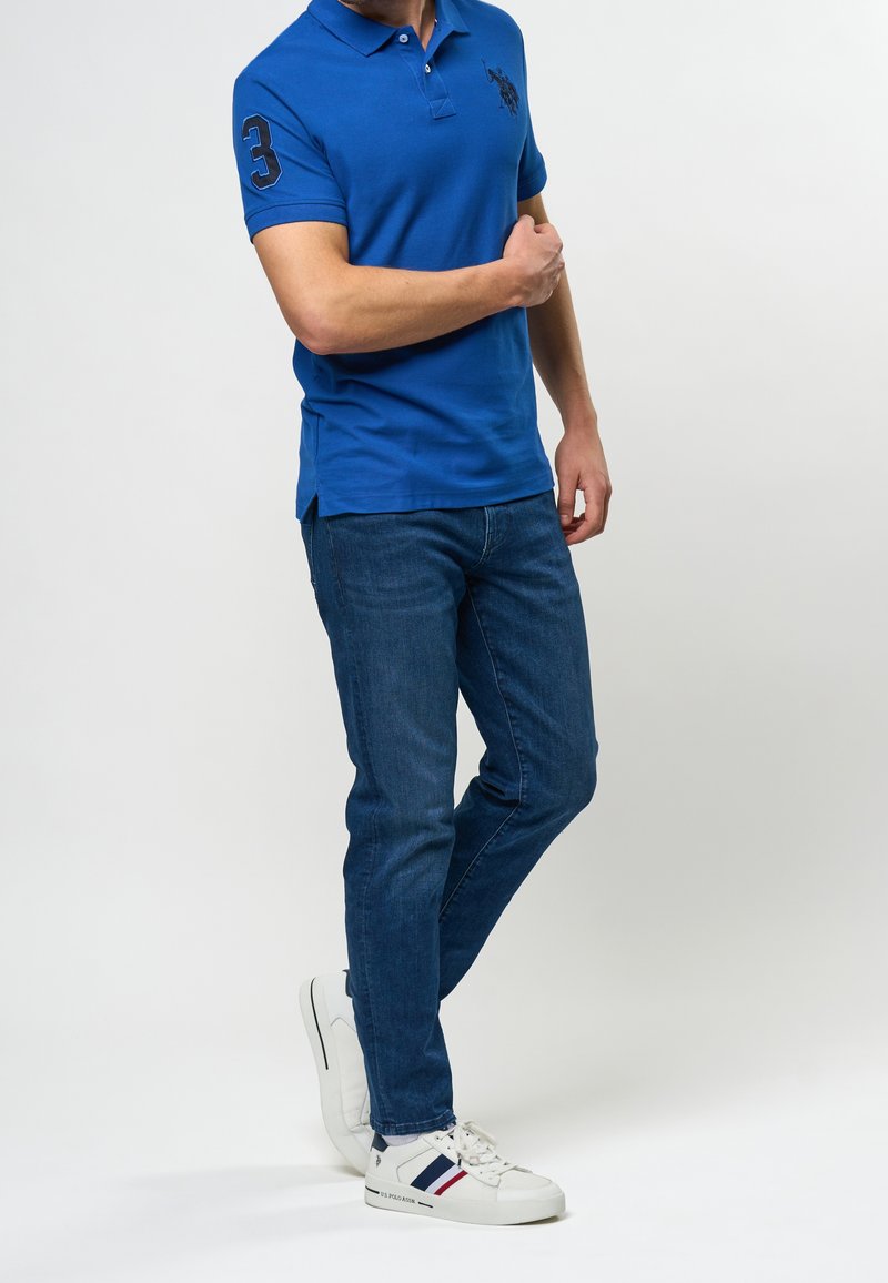 Homme portant un polo bleu, un jean bleu foncé et des baskets blanches avec des rayures rouges et bleues, posant sur un fond blanc.