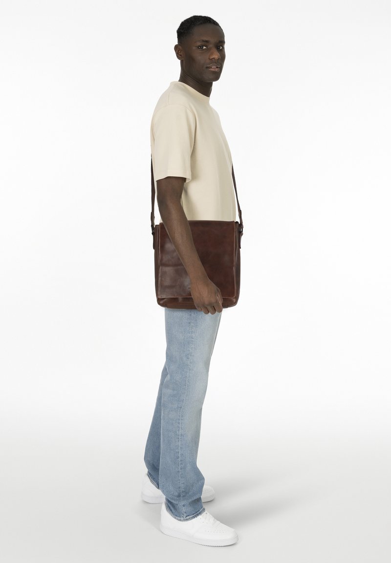 Borsa a tracolla in pelle marrone con una texture liscia, tracolla regolabile e una forma rettangolare semplice. Indossata da una persona in una maglietta chiara.
