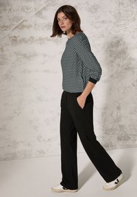 Pantaloni neri a gamba larga abbinati a un maglione a righe geometriche verde e bianco. Il modello indossa sneakers bianche con dettagli neri.