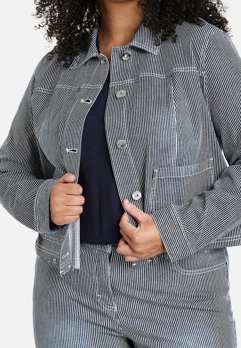 Vrouw die een opengeknipte blauw-wit gestreepte jas en bijpassende broek aanpast, over een donker topje.