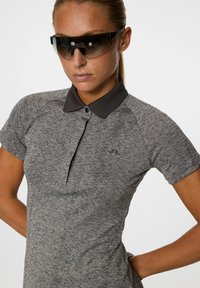 Vrouw in donkere wraparound-zonnebril en een grijze polo met korte mouwen en een donkere kraag, staand met handen op de heupen tegen een witte achtergrond.