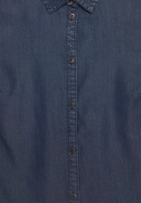 Jeansskjorta i mörkblått med en mjuk textur, försedda med en spetsig krage och sex svarta knappar som löper längs framsidan.