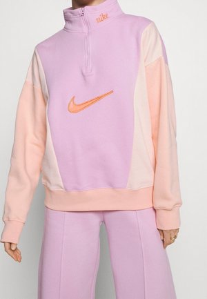 Vrouw draagt een pastelroze en paarse Nike fleecetrui met 1/4 ritssluiting en bijpassende broek, met een oranje Nike swoosh-logo op de voorkant.