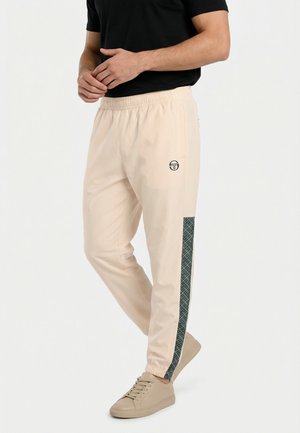Uomo che indossa pantaloni jogger beige con una striscia laterale scura a motivo e sneakers beige, in piedi contro uno sfondo chiaro uniforme.