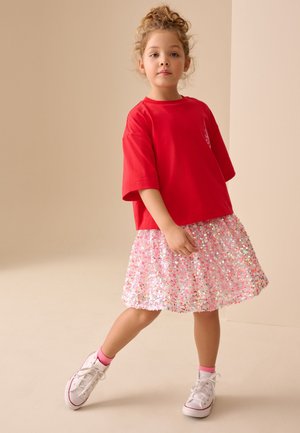 Młoda dziewczynka o kręconych włosach, ubrana w czerwoną, oversize'ową koszulkę, różową cekinową spódnicę, białe trampki i różowe skarpetki, stojąca na jednej nodze.