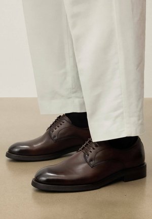 Vīriešu tumši brūnas ādas kurpes ar šņorēm, valkātas kopā ar melnām zeķēm un gaiši bēšām biksēm, stāv uz gludas grīdas.