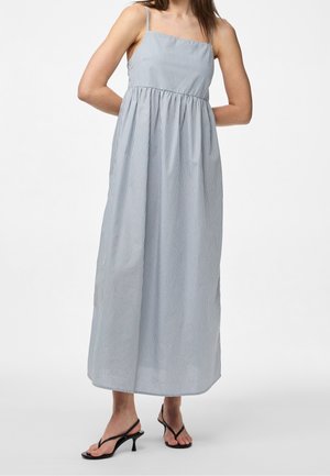 Kobieta ubrana w niebieską, bezrękawnikową, sięgającą kostek sukienkę z cienkimi pionowymi paskami i czarne sandały na obcasie.