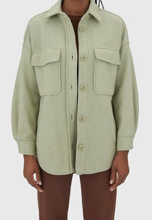 Veste boutonnée vert clair avec deux grandes poches à rabat, portée par-dessus un haut marron et associée à un pantalon marron.