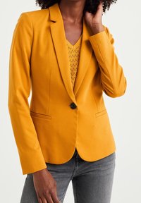 Senapsgul kavaj med notchad krage och enkel knappstängning, med två framfickor och skräddarsydd passform. Bärs över en texturerad topp.