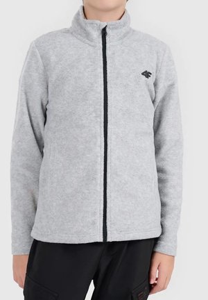 Personne portant une veste polaire zippée gris clair avec une fermeture éclair noire et un logo noir sur la poitrine, associée à un pantalon noir.