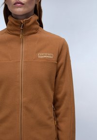 Braune Fleecejacke mit hohem Kragen, Frontreißverschluss und Logo-Patch auf der Brust. Verfügt über strukturiertes Material und passende Reißverschlussakzente.