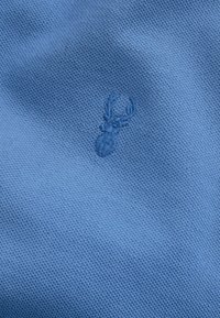 Lyseblåt struktureret stof med et broderet hjortelogo i en lidt mørkere blå nuance. Ensartet overflade med et åndbart meshmønster.
