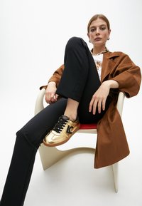 Zlaté sportovní boty s černými akcenty, s logem, nošené s černými kalhotami a hnědým zamšovým kabátem, sedící na židli s červeným sedákem.