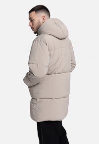 Jachetă pufoasă de culoare gri deschis, cu glugă, textură matlasată, croială lejeră și manșete elastice. Design simplu, fără logo-uri vizibile.