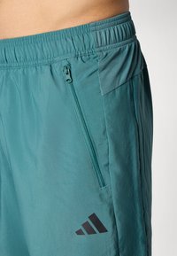 Pogled od blizu na teal atletske kratke hlače z elastičnim pasom, zadrgo na stranski žep, mrežno ploščo in črn Adidasov logotip blizu robu.