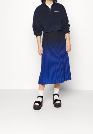 Personne portant un sweat Adidas court noir, une jupe plissée bleue et noire, des chaussettes blanches et des sandales plateformes noires, debout devant un fond blanc.