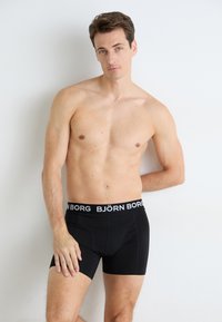 Oberkörperfreier Mann mit kurzen braunen Haaren, der schwarze Björn Borg Boxershorts trägt und an einer schlichten weißen Wand steht, mit einer Hand auf dem Oberschenkel.