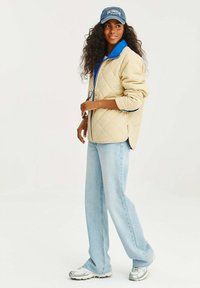 Béžová prošívaná bunda s modrými akcenty na límci, světlé modré džíny a stříbrné tenisky. Šedá čepice s nápisem "LOS ANGELES". Žádné viditelné vzory.