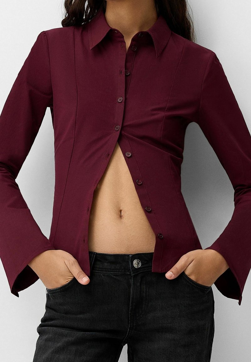 Bordeaux knäppskjorta med en figurnära design, långa ärmar och synliga sömmar, tillsammans med svarta jeans med en framficka.