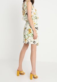 Robe courte fleurie en tissu blanc, ornée de motifs jaunes et verts. Stylée avec des sandales à talons blocs jaunes. Design sans manches.