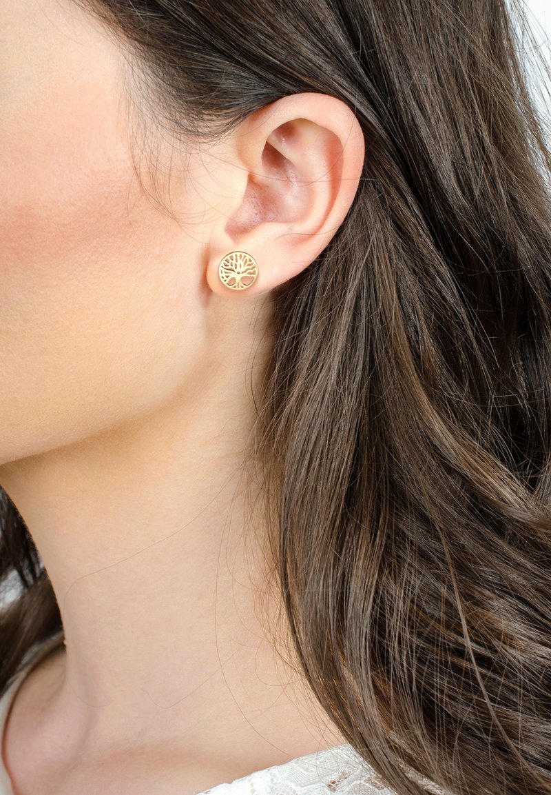 Goldene, kreisförmige Ohrringe mit einem Baumdesign-Ausschnitt. Die glatte Oberfläche und das zarte Muster stehen im Kontrast zu dunkel gewelltem Haar.