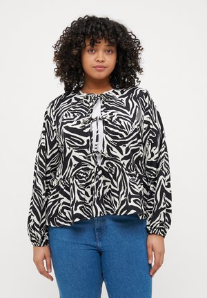 Blouse noire et blanche à motifs avec des manches longues, un design péplum et une fermeture par lien au niveau du cou. Associée à un jean bleu.