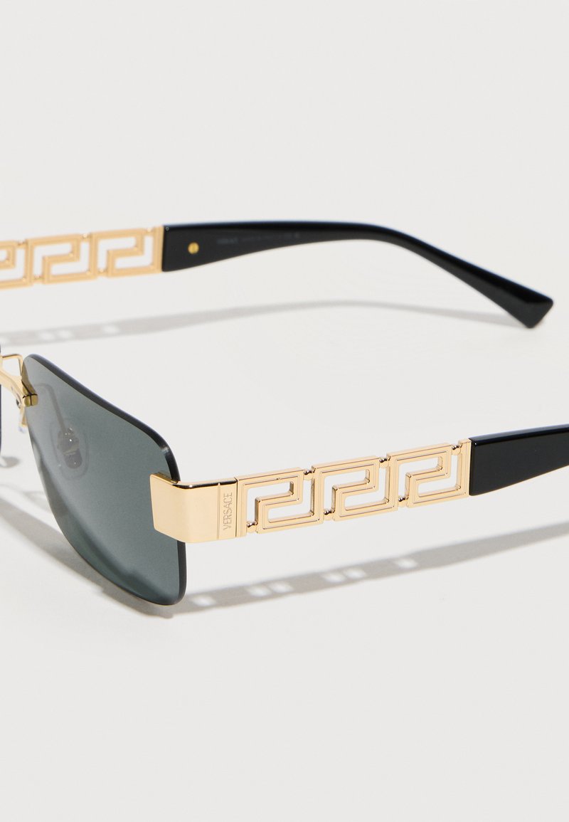 Gold and black sunglasses with dark lenses, featuring a decorative Greek key pattern on the temples and "Versace" engraved on the hinge.