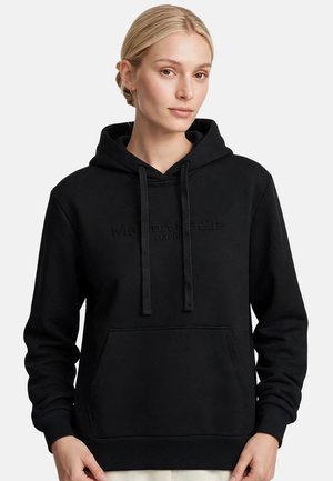 Mujer rubia con el cabello recogido en un moño bajo lleva una sudadera con capucha negra con bolsillo delantero y cordones, mirando ligeramente hacia un lado.