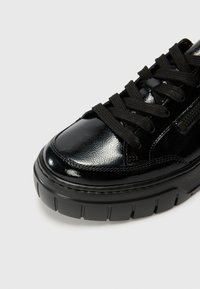 Sneaker en cuir verni noir avec un bout rond, une semelle épaisse noire et des lacets plats noirs. Comprend un détail de fermeture éclair sur le côté pour plus de fonctionnalité.