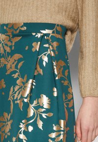 Jupe turquoise avec motif floral doré, associée à un pull en tricot côtelé beige, montrant la taille et le bras droit de la personne portant le vêtement sur fond gris.