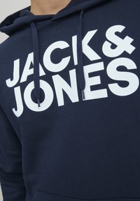 Tmavě modrá mikina s kapucí vyrobená z bavlněné směsi, s velkým bílým nápisem "JACK & JONES" na přední straně, nastavitelnou kapucí se šňůrkou a přední kapsou.