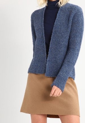 Femme portant un cardigan bleu texturé ouvert devant, un col roulé marine et une jupe couleur camel, debout devant un fond clair uni.