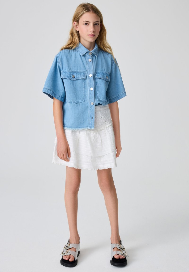 Lyseblå denim kortærmet skjorte med to forlommer og sølvknapper, kombineret med en hvid lagdelt nederdel og sorte sandaler.