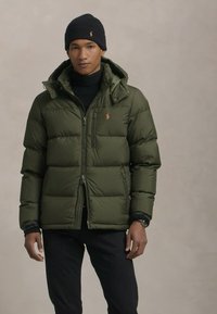 Olijfgroene pufferjack met rits, van nylon materiaal en een afneembare capuchon. Voorzien van een logo, ritszakken en geribbelde manchetten.