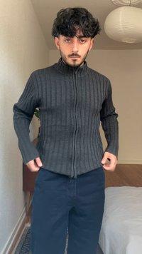 Suéter gris con cremallera, de canalé y con cuello alto, de mangas largas y diseño ajustado. Lleva puesto sobre pantalones oscuros en un entorno interior luminoso.