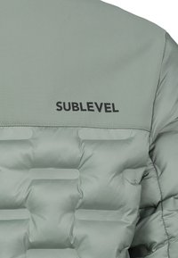 Hellgrüne gepolsterte Jacke mit Rückendetail, das mit gestickten rechteckigen Mustern und dem schwarzen "SUBLEVEL"-Logo-Text versehen ist.