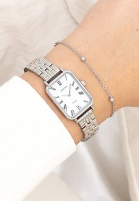 Montre-bracelet en argent avec un cadran rectangulaire, un fond blanc, des chiffres romains noirs, et un bracelet en maillons métalliques. Un délicat bracelet en chaîne argentée l'accompagne.