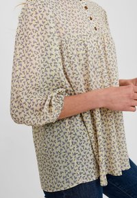 Blouse jaune clair avec un motif floral en violet et vert. Présente un col rond, des boutons et des manches élastiques froncées.