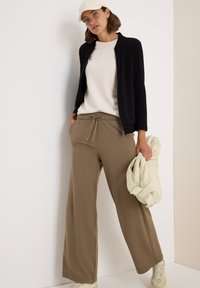 Veste zippée noire, pull beige et pantalon taupe à jambes larges. Le modèle tient un sac vert clair. Une casquette et des baskets claires complètent la tenue.