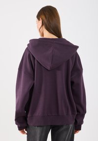 Moletom roxo oversized feito de mistura de algodão, apresentando um grande capuz, ombros caídos e punhos canelados. O tecido parece suave.