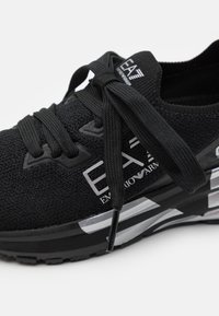 Černá atletická bota s pletenou texturou, s logem a stříbrnými akcenty. Obsahuje ploché černé tkaničky a polstrovanou podrážku.