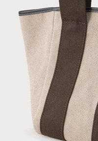 Καμβάς τσάντα χιαστί σε μπεζ απόχρωση με υφή, διαθέτοντας σκούρο καφέ ύφασμα για λουρί και αντίθετη κυρτή άκρη στην κορυφή.