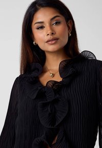 Czarna plisowana bluzka z falbankowatym dekoltem i rękawami. Tkanina ma teksturowane wykończenie, a subtelny połysk dodaje głębi.