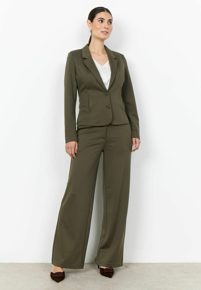 Femme debout portant un blazer vert olive et un pantalon à jambes larges avec un haut blanc et des chaussures marron pointues contre un mur blanc.