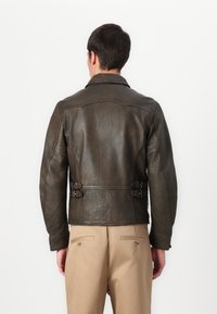 Veste en cuir marron avec une silhouette structurée, une texture subtile et des accents de boucle à la taille. Elle présente un col classique et une coupe ajustée.