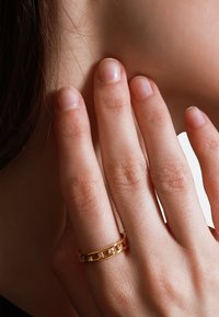 Gouden ring met verstrengelde schakelshape, met een gladde textuur en gepolijste afwerking. Draagt op een uitgestoken vinger, waardoor het ontwerp en de vorm benadrukt worden.