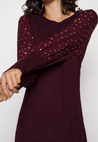 Robe bordeaux à manches longues, avec un tissu texturé et des motifs dorés sur les manches, des poignets côtelés et un col rond classique.